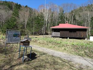 青空のもと、赤い屋根の北稜岳ヒュッテと入林届ボックスの風景が、自然の中に溶け込んでいます。