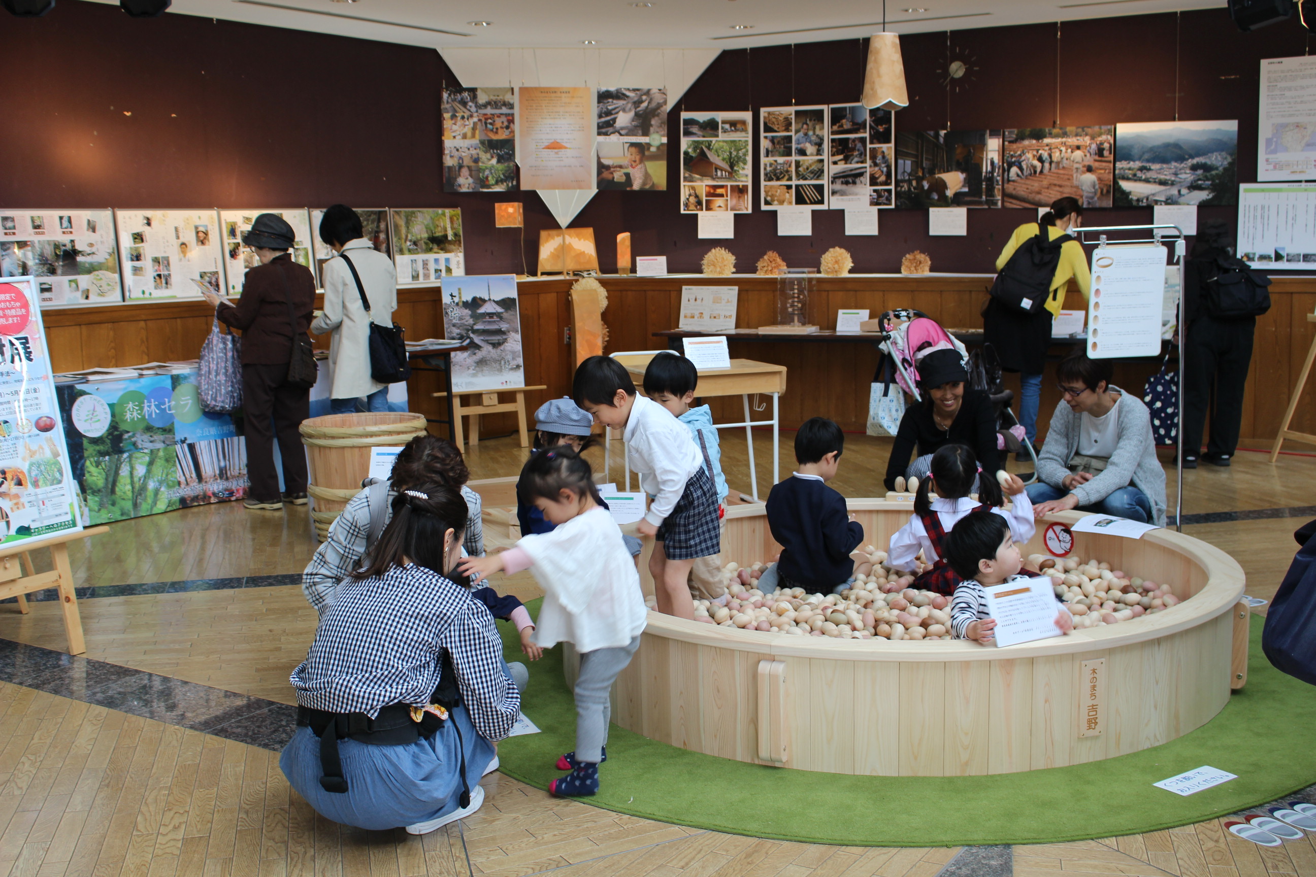 木の球プールで遊ぶ子供達と展示を見学する人達