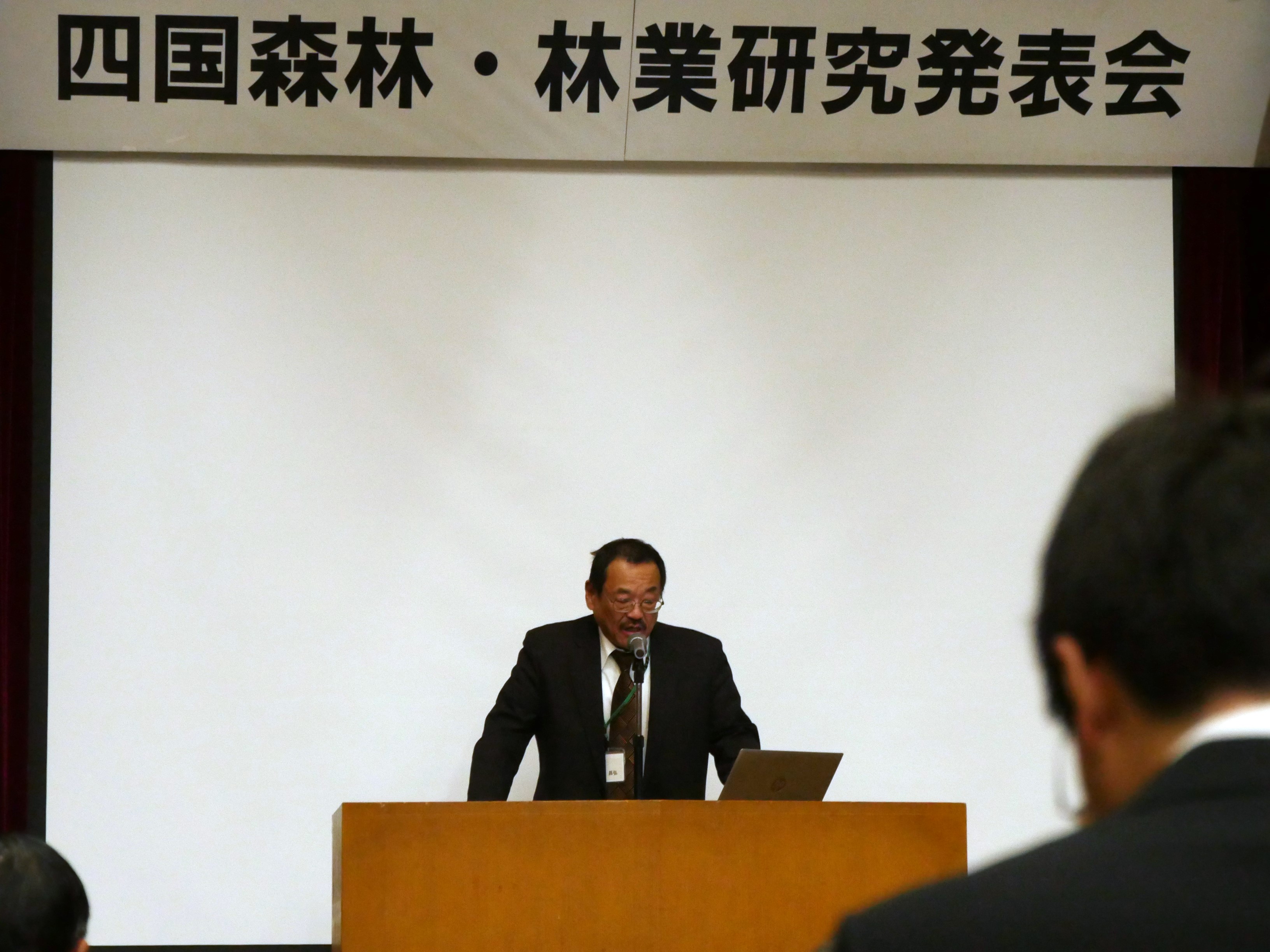 審査委員長が、ステージ中央で演台の後ろに立ち、講評を述べる様子