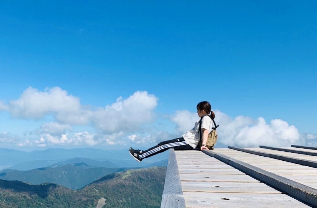 青空の下、雲を見下ろすような高所に設けられた木製のデッキに座る人物が印象的です。視界いっぱいに広がる澄んだ青と白い雲が、開放感と非日常の空気を演出しています。初めての登山で自然と一体になり、静かな時間を楽しむ様子が伝わる写真です。