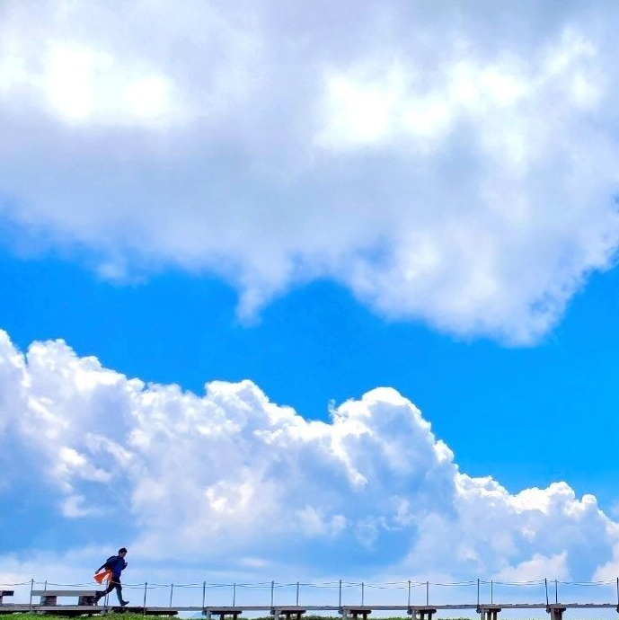 澄み渡る青空に、白くふわりと広がる雲が印象的な一枚です。画面下には細い橋が伸び、その上を走る人の姿が小さく映っています。広大な空と雲のスケール感に対し、人の存在が控えめで、自然の雄大さと日常の対比が際立つ風景です。