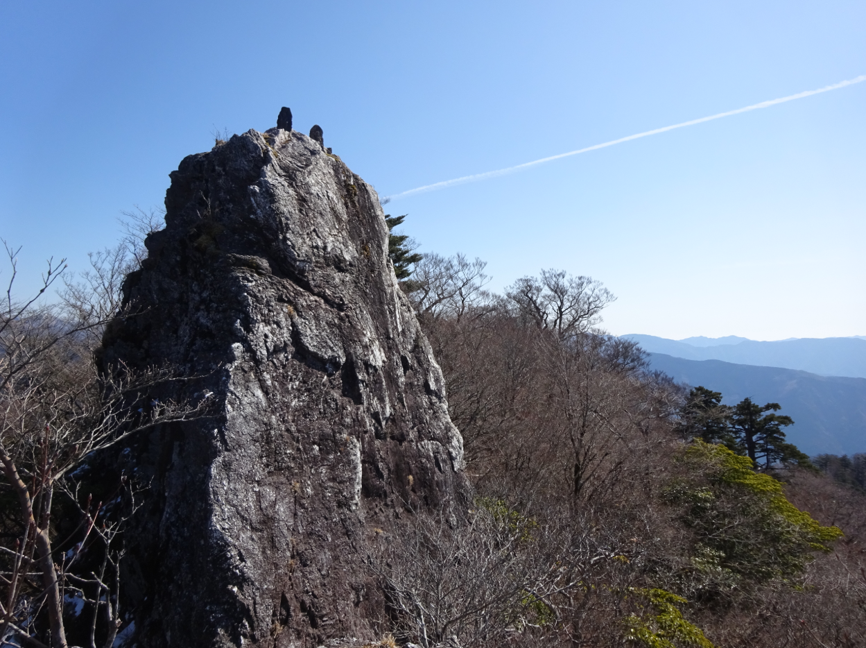 岩肌がむき出しになった大きな岩峰が、周囲の山並みの中で際立っています。背景には遠くの山々が重なり、澄んだ青空に一本の飛行機雲が伸びて、静けさと広がりを感じさせる風景です。