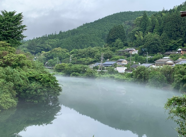 川面に淡い霧が立ち込め、静かな朝の雰囲気を漂わせています。周囲には濃い緑の木々が広がり、奥には山の斜面と集落の家々が見え、自然と人の暮らしが調和した風景です。水面は鏡のように穏やかで、霧が柔らかく景色を包み込み、幻想的な印象を与えています。