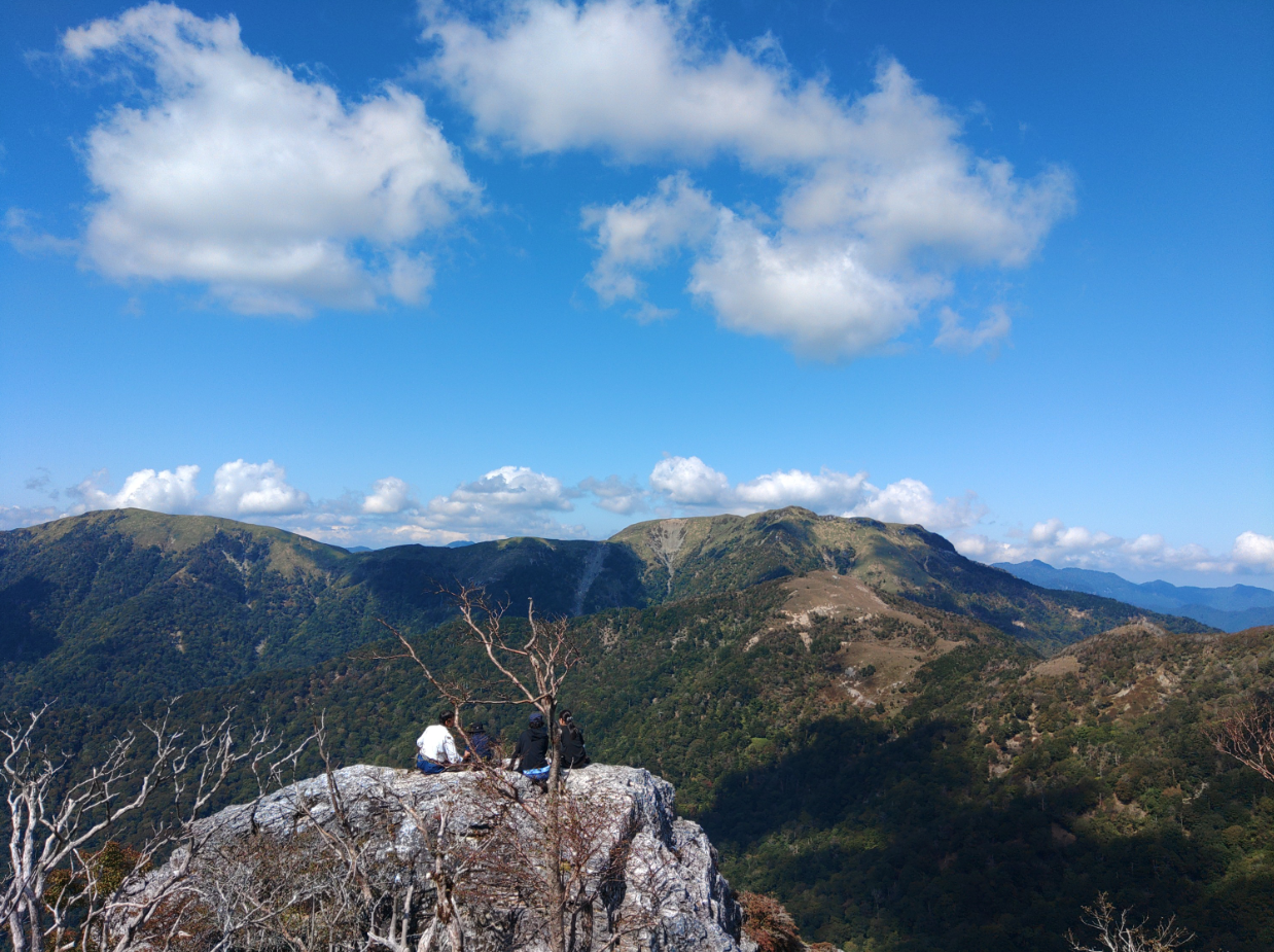 岩場の上から広がる雄大な山並みを見渡す風景です。前景には岩の上に座る人物の後ろ姿があり、その先には緑と茶色が入り混じる山々が連なっています。青空には白い雲が浮かび、澄んだ空気と広がりのある景観が印象的です。