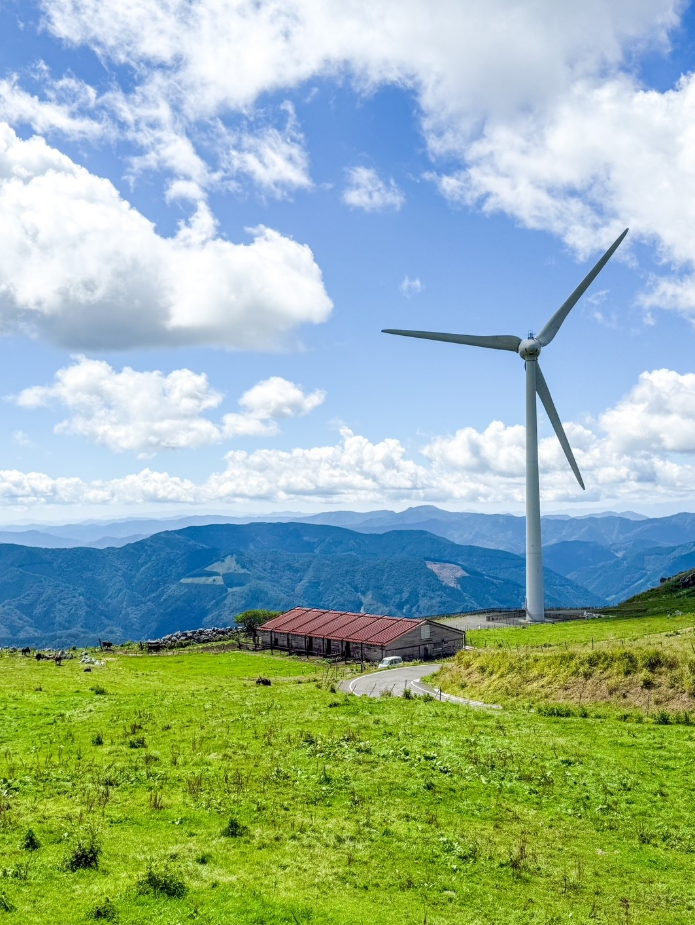 青空の下、広々とした草原に大型の風力発電機が立っています。前景には緑の牧草地と赤い屋根の建物があり、背景には連なる山々が遠くまで続いています。