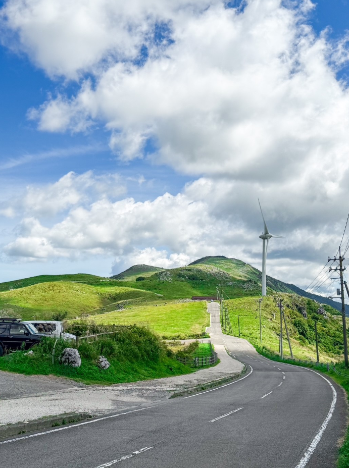青空と白い雲が広がる中、緩やかな丘陵地を走る舗装道路が印象的です。道路脇には草原が広がり、鮮やかな緑が風景を彩っています。遠くには小高い山が見え、自然の雄大さと開放感を感じる景色です。