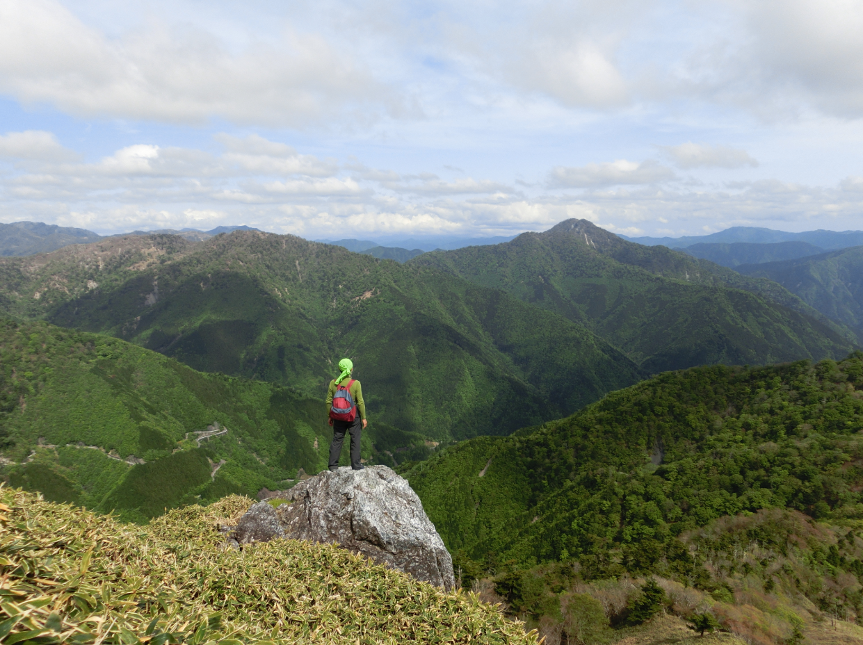 岩場に立つ登山者の背中越しに、連なる深緑の山々が広がる雄大な景色です。遠くまで続く稜線と、雲が浮かぶ穏やかな空が、自然の広がりと静けさを感じさせます。